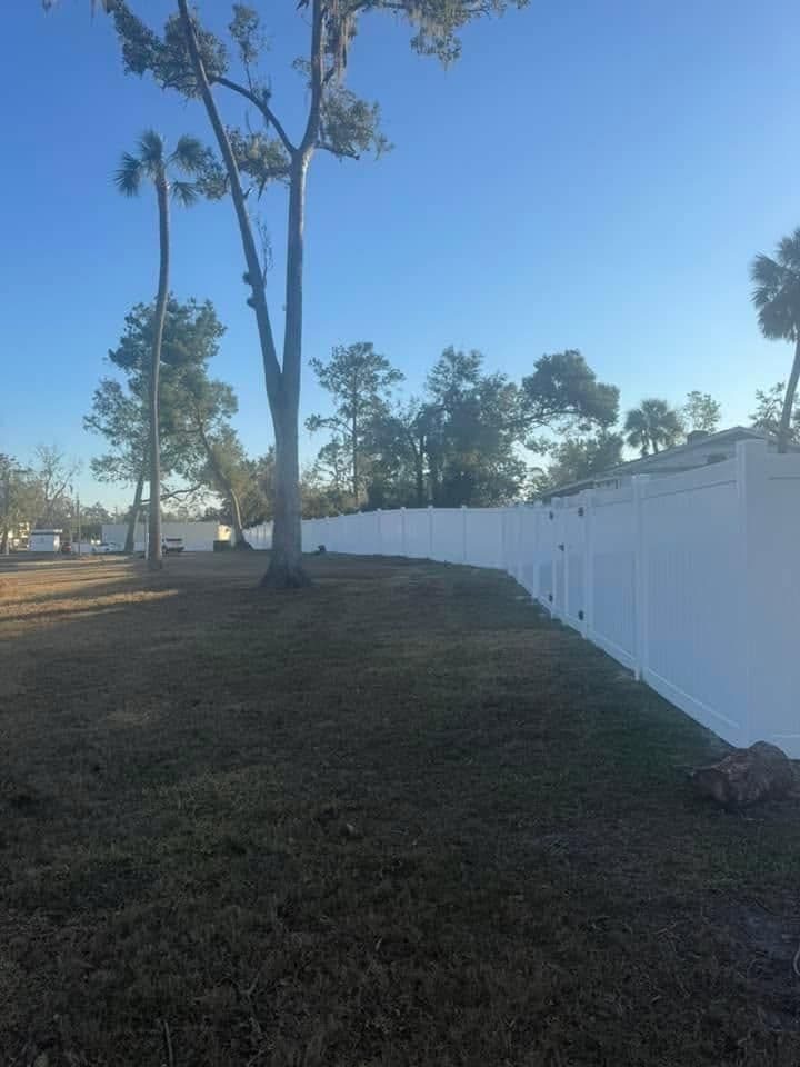 White vinyl fence along a grassy area with tall trees under a clear blue sky.