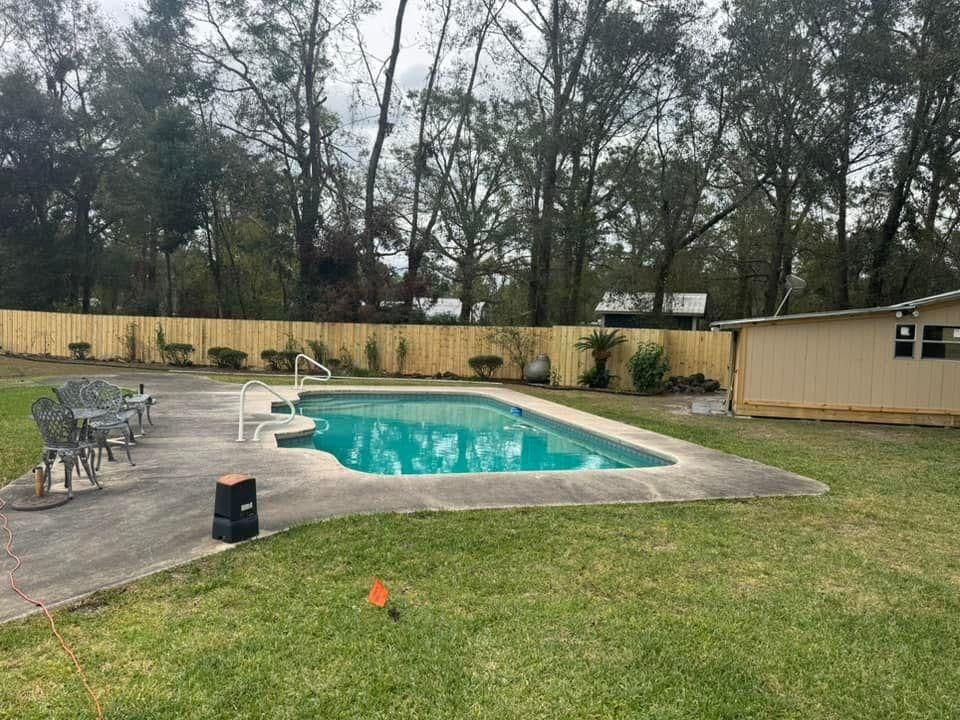 Backyard with swimming pool, patio chairs, and fenced greenery under cloudy sky.