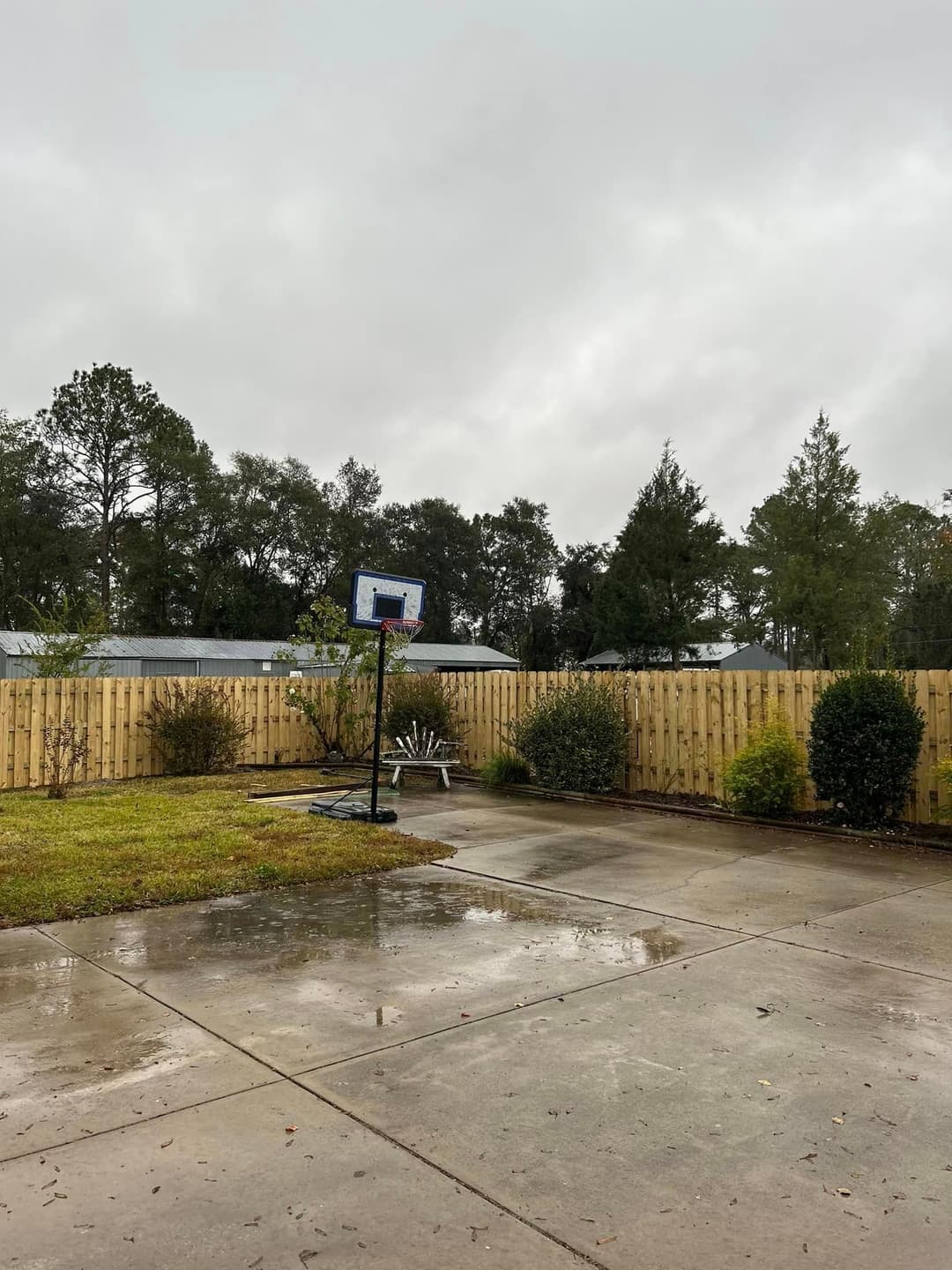 Basketball hoop on wet pavement in a backyard with cloudy sky and wooden fence.