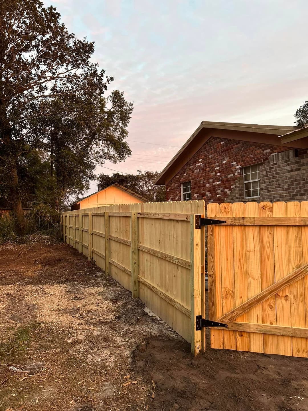 New wooden fence with gate separating a yard from a brick house at sunset.