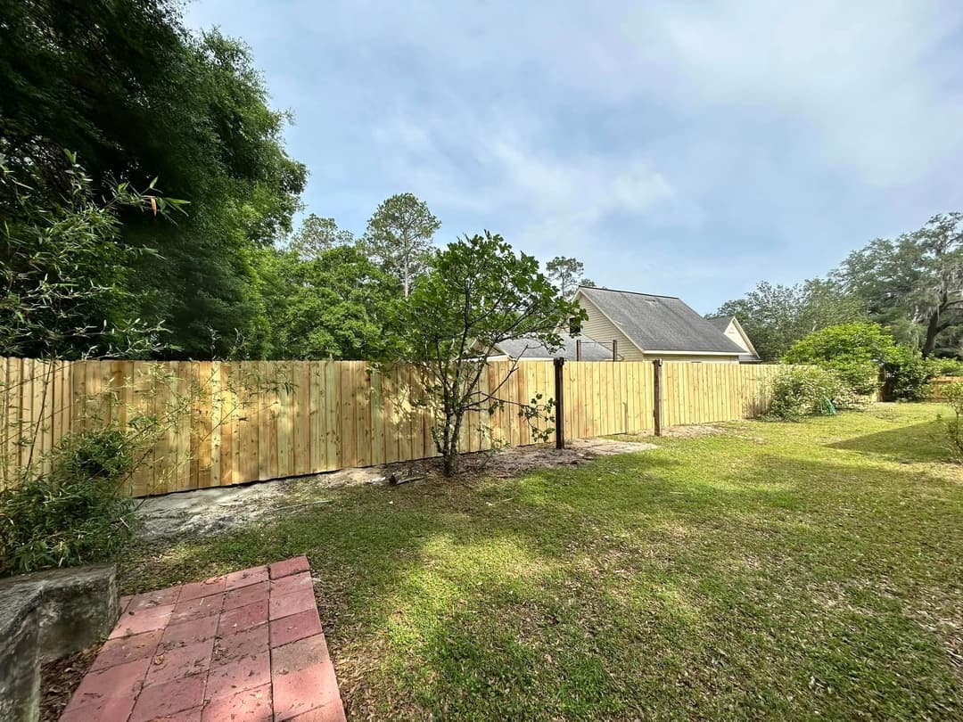 Wooden fence enclosing a backyard with green grass and trees under a cloudy sky.