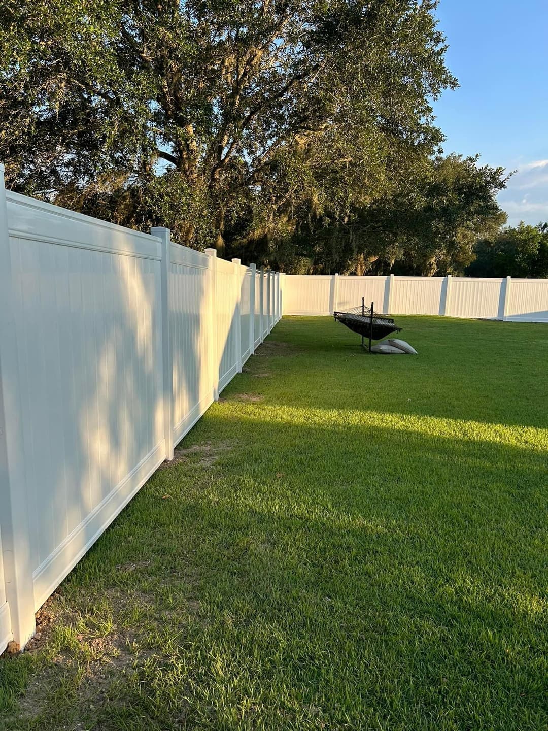 White vinyl fence along a green lawn with a large tree and a hammock in the background.