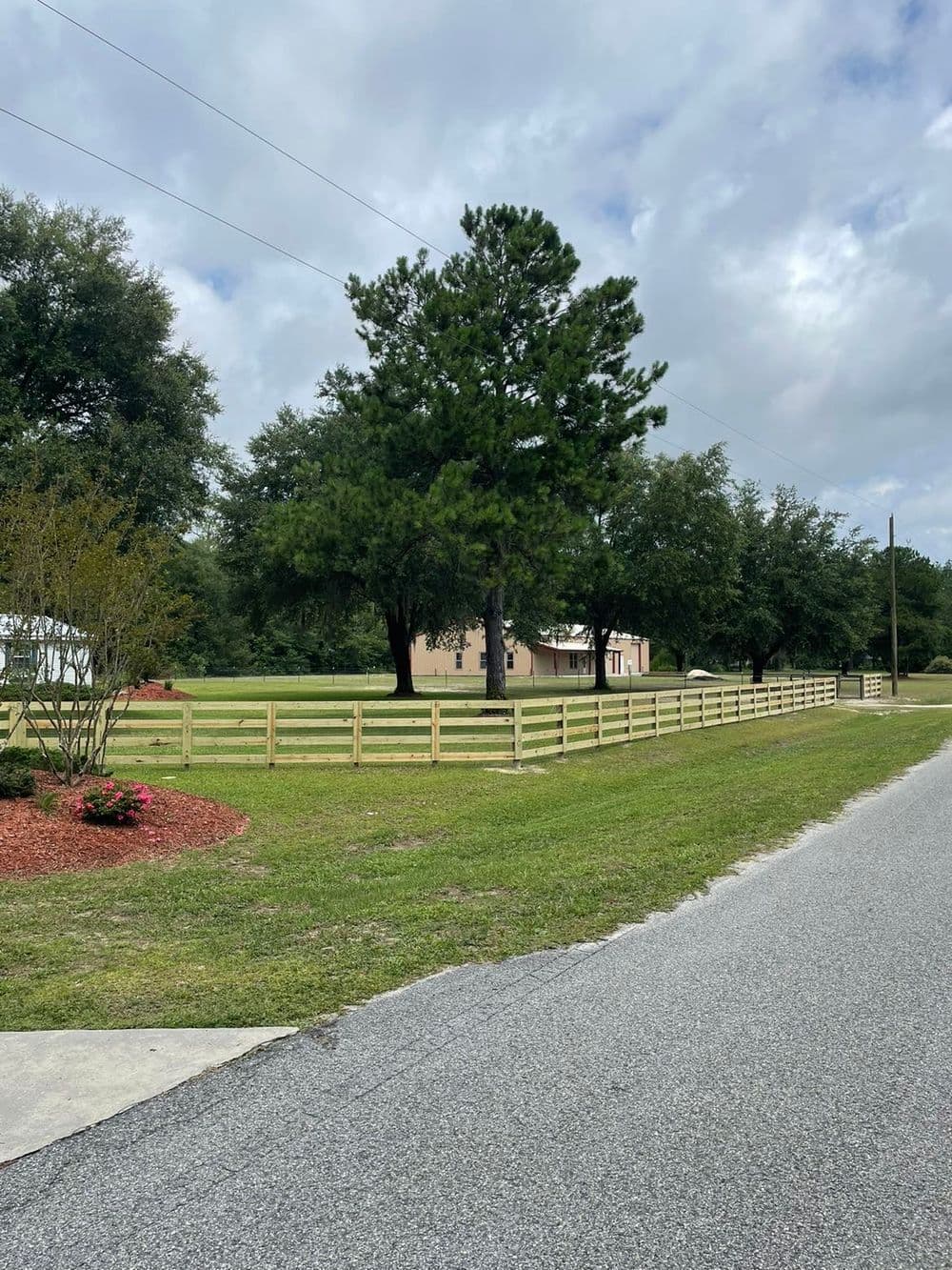 Country road view featuring a wooden fence, green trees, and a house in a rural setting.